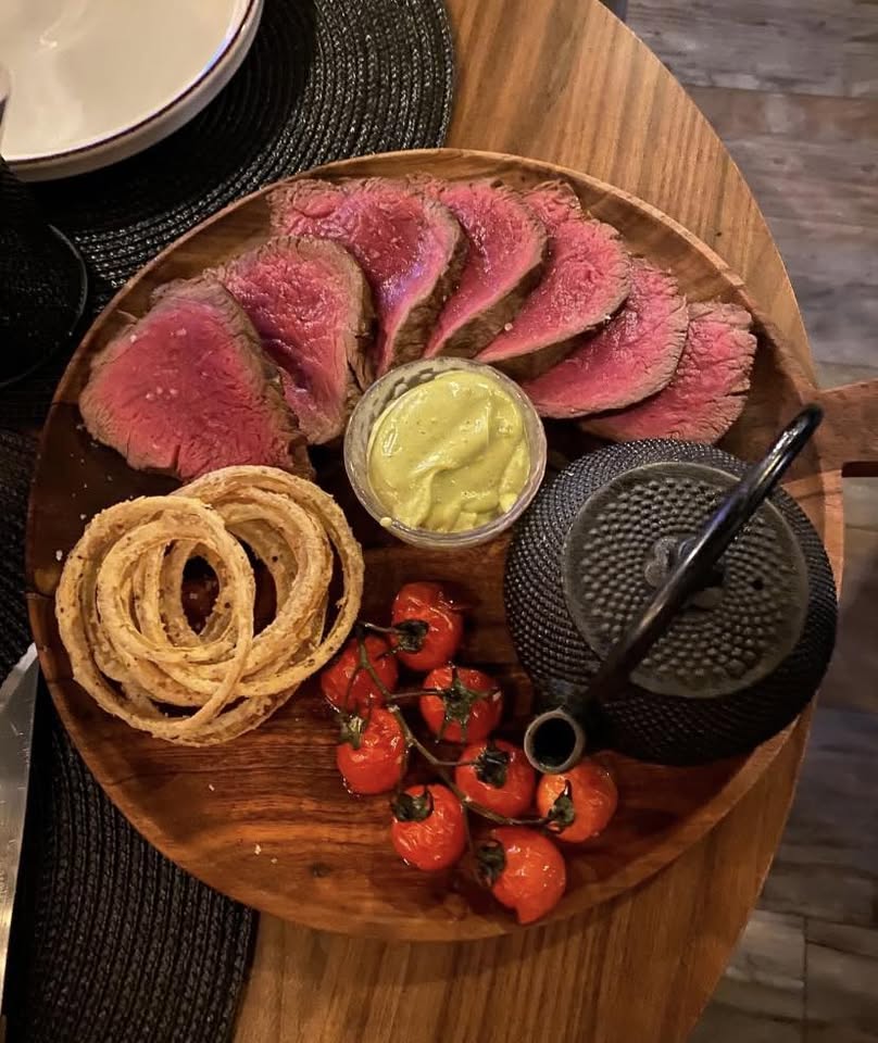 Sliced beef tenderloin served medium rare on a wooden board with mustard, onion rings and cherry tomatoes